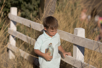 a child in a T-shirt walks near a white wooden fence and holds on to it with one hand, and in the other hand carries his glass bottle
