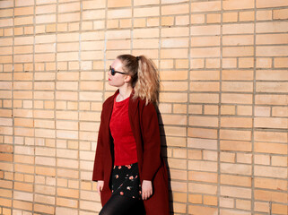 Young blond woman wearing sunglasses, woolen coat, red sweater and floral print skirt is standing...