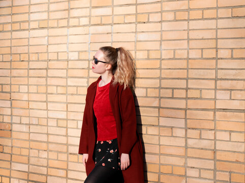 Young blond woman wearing sunglasses, woolen coat, red sweater and floral print skirt is standing by the brick wall on a sunny day. Spring street style