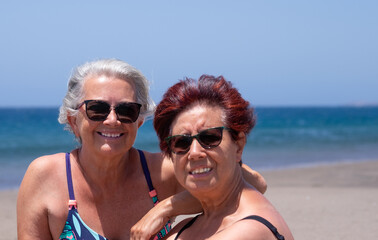 Portrait of two smiling senior women sitting on the beach looking into the camera. Blue sky and sea on background