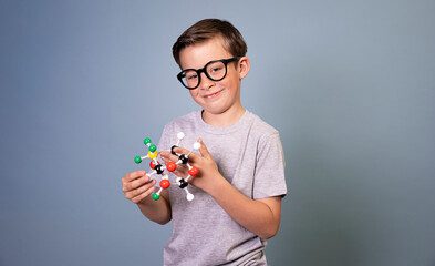 cool schoolboy with black glasses stands in front of blue background holding model of molecules