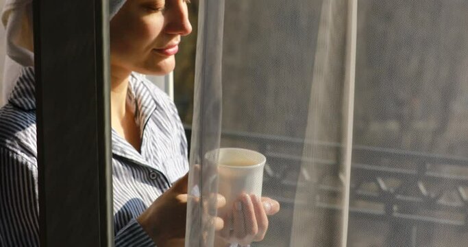 4 k. happy beautiful young woman with a towel on her head is having breakfast on the balcony, drinking coffee or tea. girl looks out over the rooftops of the city morning time