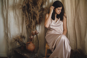 Stylish woman in silk dress sensually posing in boho room with dry grass. Modern bohemian bride