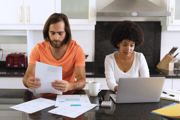 Diverse couple sitting in kitchen using laptop and paying bills