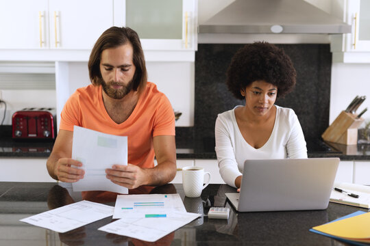 Diverse Couple Sitting In Kitchen Using Laptop And Paying Bills