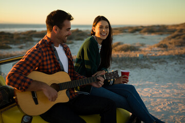Happy caucasian couple sitting on beach buggy by the sea playing guitar and drinking during sunset