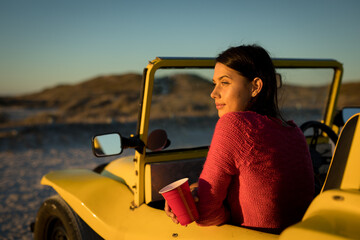 Happy caucasian woman sitting in beach buggy by the sea holding red cup