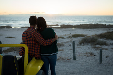 Happy caucasian couple leaning against beach buggy by the sea during sunset embracing