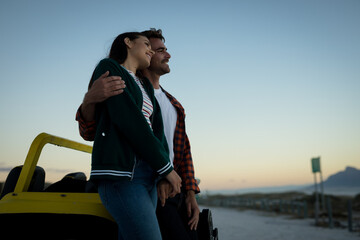 Happy caucasian couple leaning against beach buggy by the sea during sunset embracing