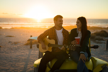 Happy caucasian couple sitting on beach buggy by the sea playing guitar and drinking during sunset