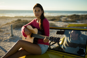 Caucasian woman sitting on beach buggy playing guitar, man sitting behind steering wheel