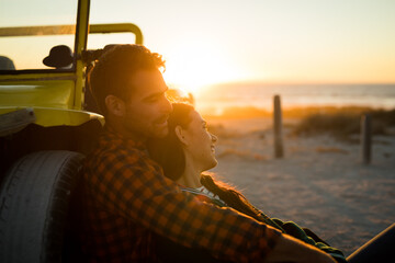 Happy caucasian couple leaning against beach buggy by the sea relaxing during sunset
