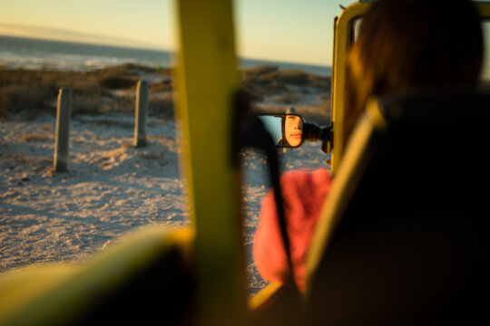 Caucasian woman sitting in beach buggy by the sea during sunset reflected in mirror - Powered by Adobe