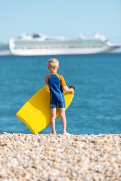 A young blonde boy standing on a rocky beach looking towards the sea holding his bodyboard. The photo is taken in Weymouth during summer. There is a cruise ship out of focus in the background.
