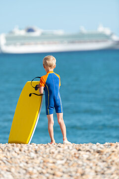 A young blonde boy standing on a rocky beach looking towards the sea holding his bodyboard. The photo is taken in Weymouth during summer. There is a cruise ship out of focus in the background.