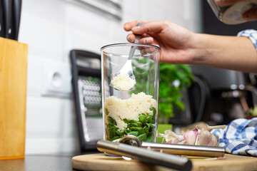 The person adding the grated cheese to the blending container. Preparation of pesto.