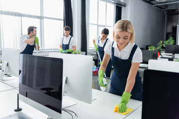 Smiling cleaner cleaning table with detergent near multiethnic colleagues and computers in office