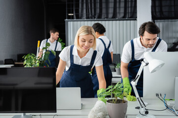 Positive cleaner holding dust brush near computers and colleague in office