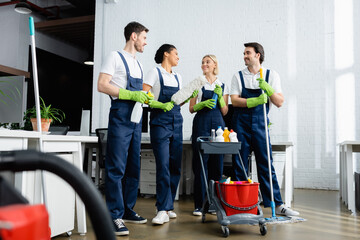 Smiling multiethnic cleaners talking near cart with detergents in office