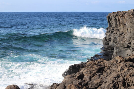 Foamy waves crashing against the brown rocks. Side view of the turquoise ocean rugh waves and blue sky. Ocean water and rocks formed from solidified lava of volcano.