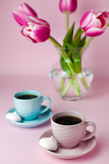 Fototapeta na wymiar A two cups of coffee and a glass vase with pink tulips on the pink background. Close-up.
