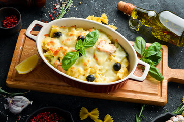 Baked farfalle pasta with salmon and cheese in a bowl on a black stone background. Top view. Rustic...