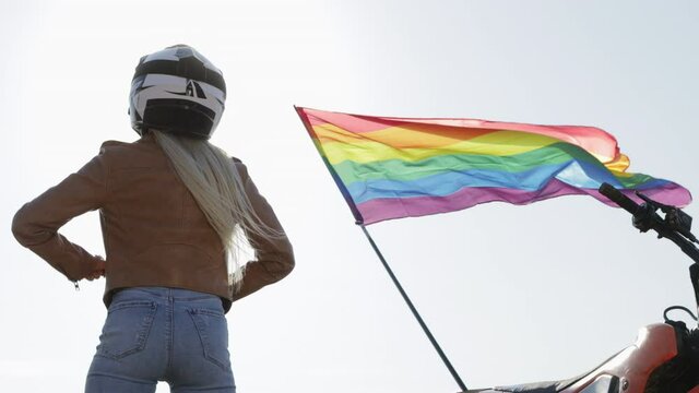 A biker girl stands in a protective helmet with her back to the camera and raises her hands up against the background of the LGBT flag waving in the wind.