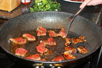 Close-up of chunks of beef frying in a frying pan. Partially view of a hand with a fork turning the beef.