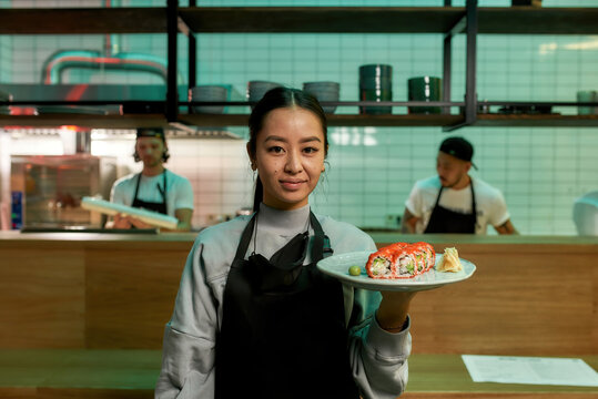 Portrait of smiling waitress wearing apron looking at camera, holding a plate with sushi rolls. Japanese cuisine in restaurant