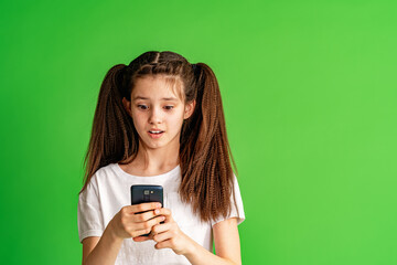 cute teenage child, looking shocked, scared, looks in amazement at a smartphone holding a cell phone in his hand on a green background in the studio. Copy space