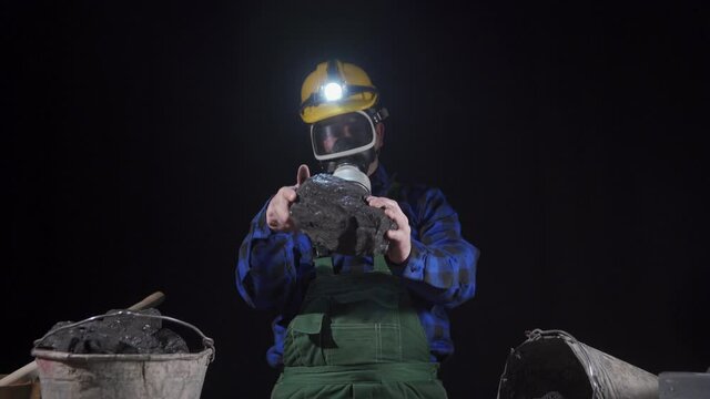 A miner wearing a dust mask and helmet with a flashlight holds a lump of black coal in his hands