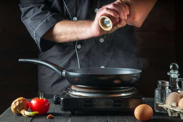 The chef adds pepper while cooking eggs in pan. Work environment on vintage kitchen table