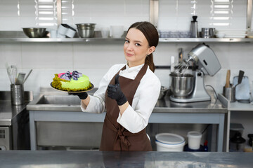 Young female chef in uniform carrying a cake tray, giving a thumbs up.