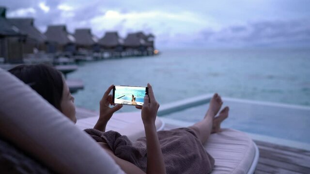 Phone - Lady using mobile cell phone social media app on vacation looking at photos posting images on social network by pool at night on travel holidays. Girl using smartphone app looking at screen.