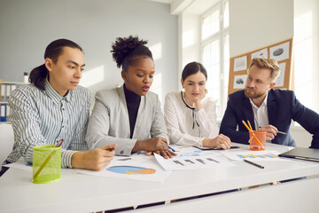 Productive teamwork in group meeting: Team of four serious smart business people sitting at office desk together, working with papers, listening to colleague, discussing further marketing strategy