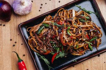 Stir fry noodles with vegetables and beef in black plate. Wooden background. Close up.