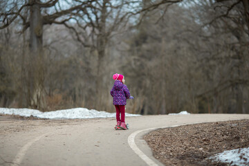 Obraz premium A European four-year-old girl in pink and purple clothes rides a scooter in a park.