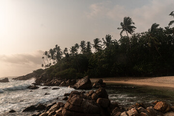 Tropikalna plaża, palmy oraz skaliste wybrzeże na tle zachodzącego słońca.