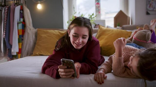 Sisters indoors at home on bed, using smartphone and having fun. Lockdown concept.