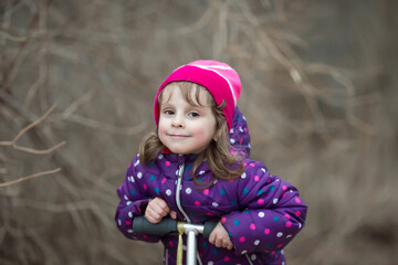 A Caucasian four-year-old girl in pink and purple clothes rides a red scooter in the park