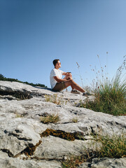 The guy dreamily looks into the distance, sitting on the stones against the background of the sky. Handsome man in sunglasses, white t-shirt and shorts.