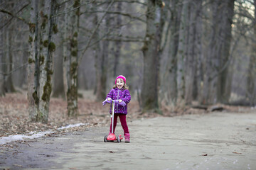 A Caucasian four-year-old girl in pink and purple clothes rides a red scooter in the park