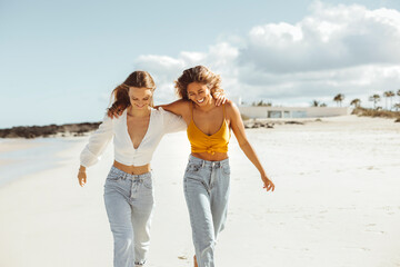 Two women walking together at the beach, hugging each other and smiling - 426158401