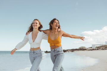 Two women walking together at the beach, hugging each other and smiling - 426158443