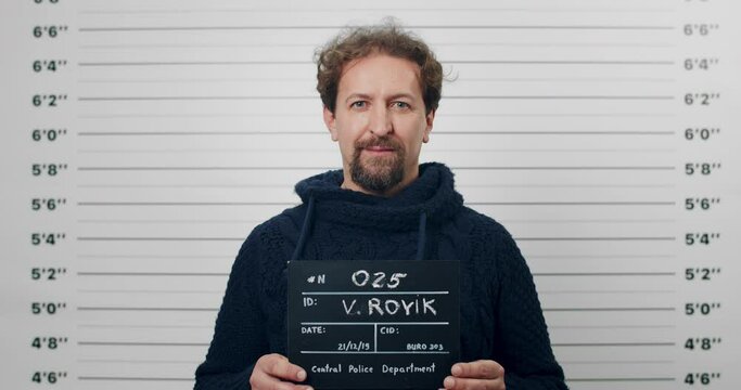Portrait of adult bearded man holding sign for photo in police department. Crop view of male person with blue eyes posing, raising head and looking to camera. Concept of mugshot and crime.
