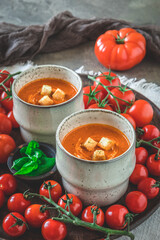Two cups filled with fresh homemade tomato soup on a wooden plate with a variety of ripe tomatoes, vertical