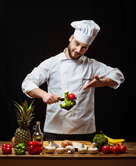 A chef in a white uniform and a cap holds a knife and vegetables
