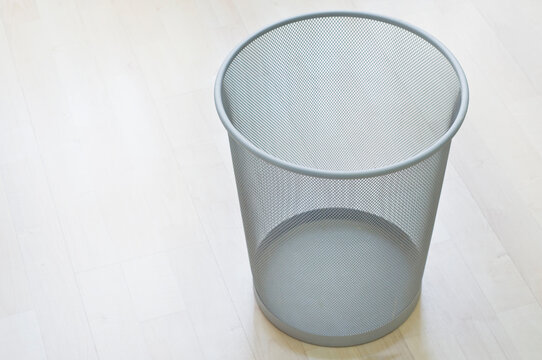 A metallic, see-through empty trash can placed on the wooden floor.