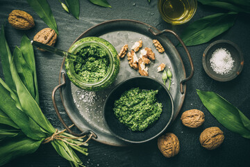 Homemade wild garlic pesto on a metal tray, wild garlic leaves, salt, oil and walnuts on dark green background, top view