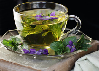 Mint or peppermint herbal tea in glass cup with fresh leaves, viola flowers on fabric on rustic background, closeup, naturopathy and aromatic spring floral teas concept